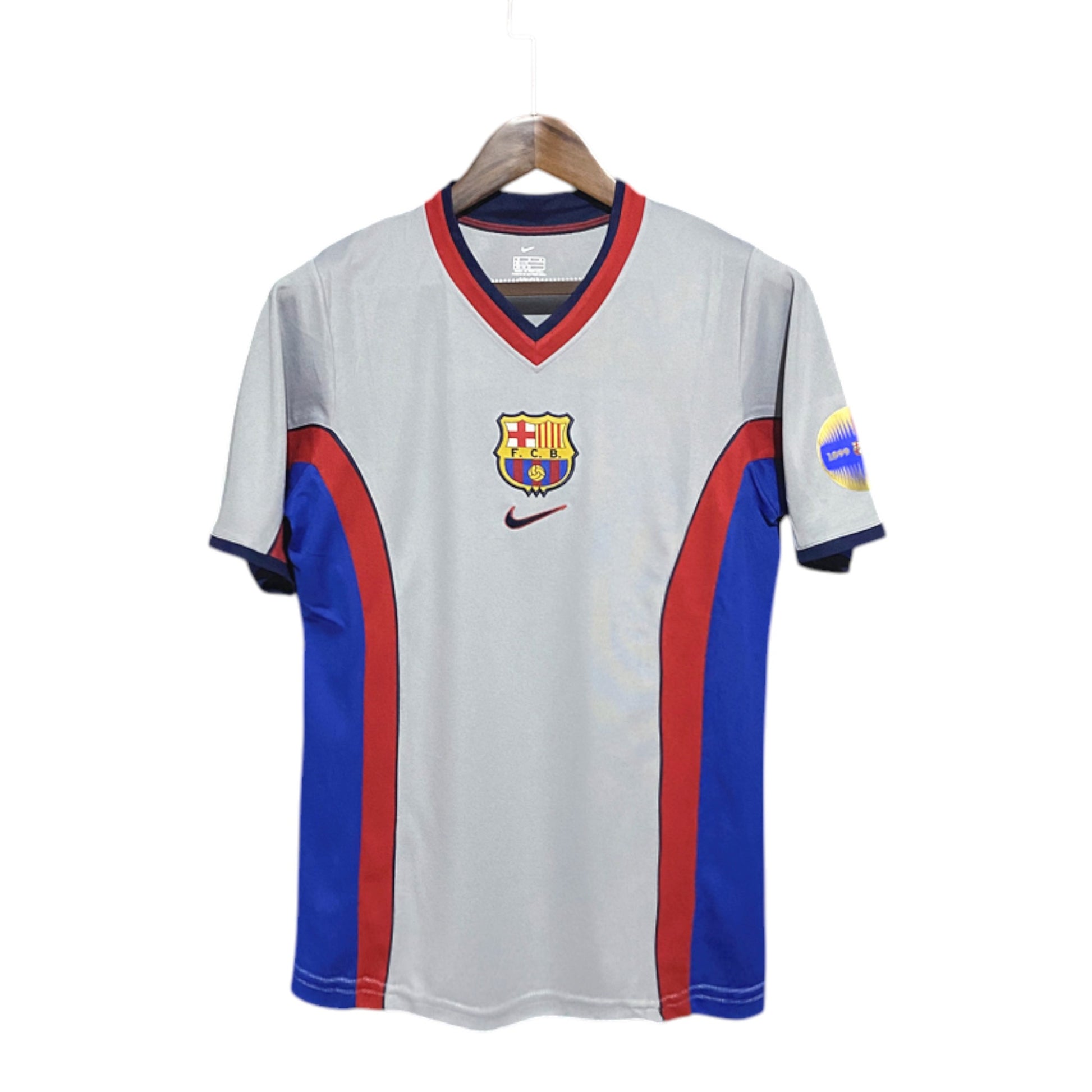 A FC Barcelona 2000/2001 Away Jersey hanging on a wooden hanger, predominantly gray with red and blue accents, and the Barcelona crest on the chest.
