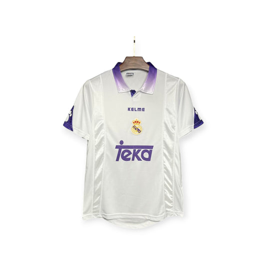 Real Madrid 1997/1998 home jersey displayed on a mannequin, predominantly white with purple accents, featuring the team's logo and a sponsor's logo.