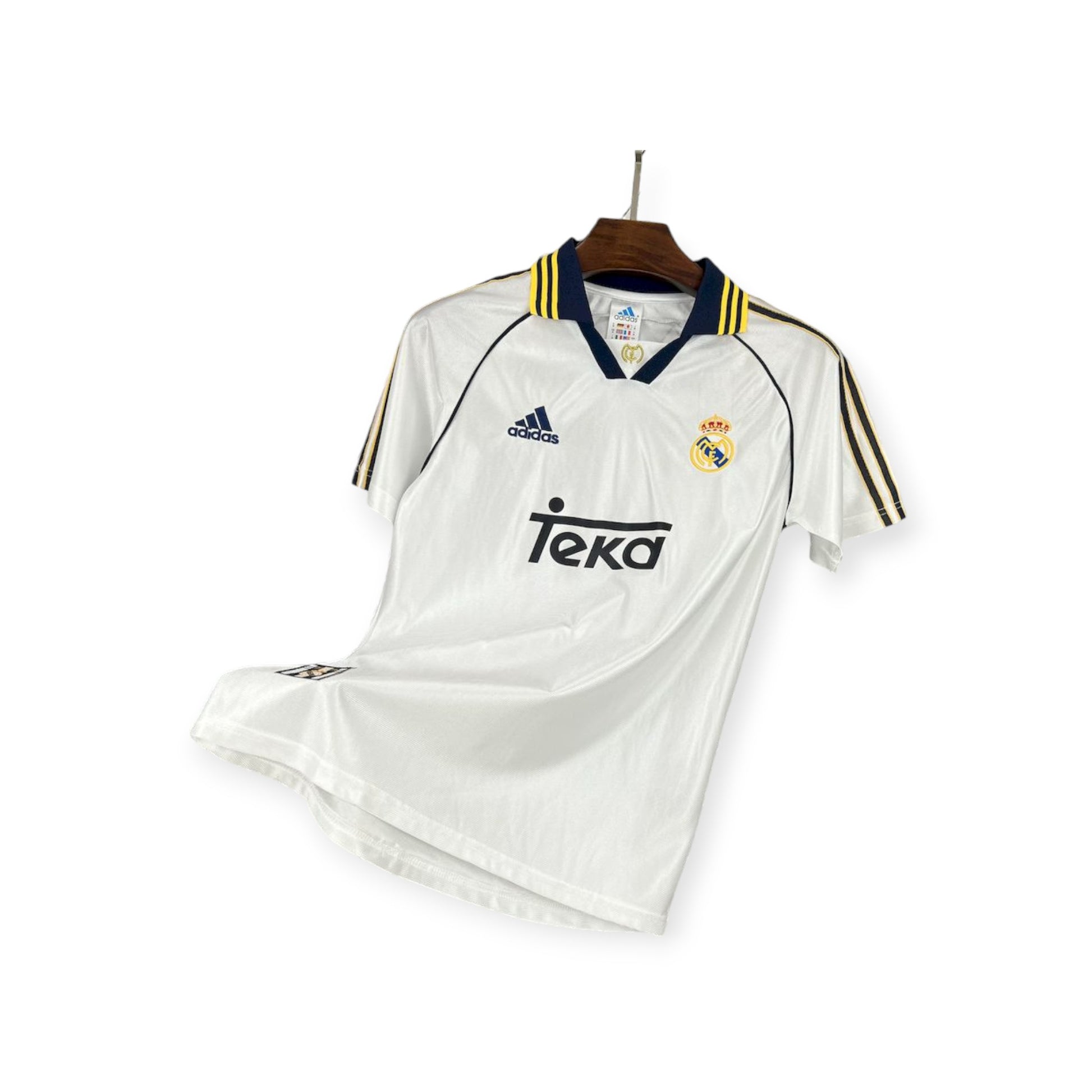 Real Madrid 1998/1999 home jersey displayed on a wooden hanger, predominantly white with yellow and blue accents, Adidas branding, and Teva sponsor logo.